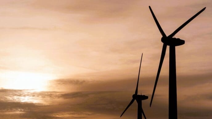Silhouettes of two wind turbines stand against a warm, cloudy sunset sky, with the sun low on the horizon.