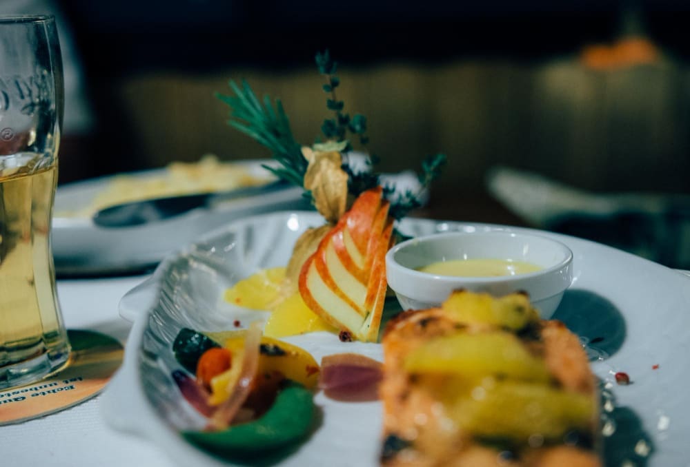 A beautifully plated dish of food, possibly a main course, is shown with a garnish of herbs and a side of sauce in a small bowl, with a glass of what appears to be beer or white wine in the background on the left.