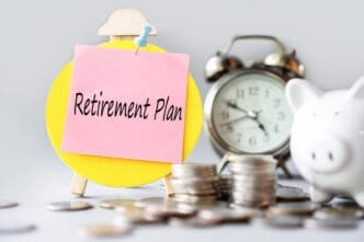 A pink sticky note with "Retirement Plan" written on it, pinned to a yellow circular easel, with a blurred piggy bank, stacks of coins, and an alarm clock in the background, symbolizing financial planning.