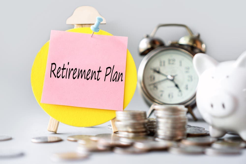 A pink sticky note with "Retirement Plan" written on it, pinned to a yellow circular easel, with a blurred piggy bank, stacks of coins, and an alarm clock in the background, symbolizing financial planning.