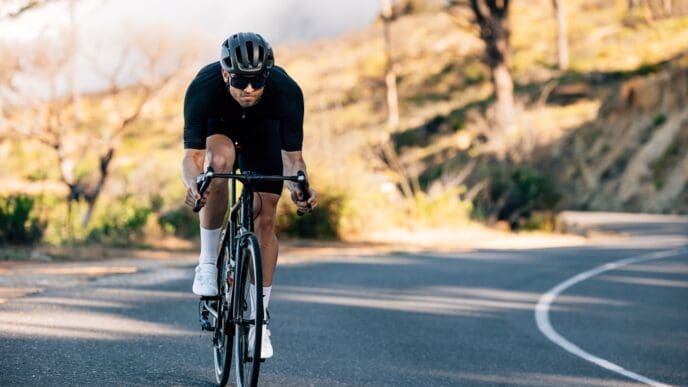 A man in black cycling gear and a helmet riding a road bicycle on a winding road with trees.