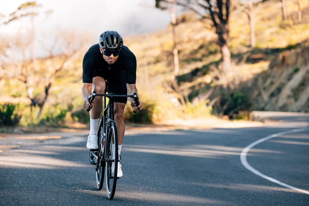 A man in black cycling gear and a helmet riding a road bicycle on a winding road with trees.