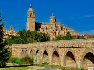 The ancient Roman Bridge of Salamanca, with its multiple stone arches, leads towards the majestic Salamanca Cathedral with its grand domes and towers, all under a brilliant blue sky, with a paved path in the foreground.