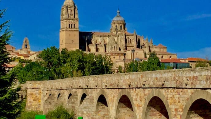 The ancient Roman Bridge of Salamanca, with its multiple stone arches, leads towards the majestic Salamanca Cathedral with its grand domes and towers, all under a brilliant blue sky, with a paved path in the foreground.
