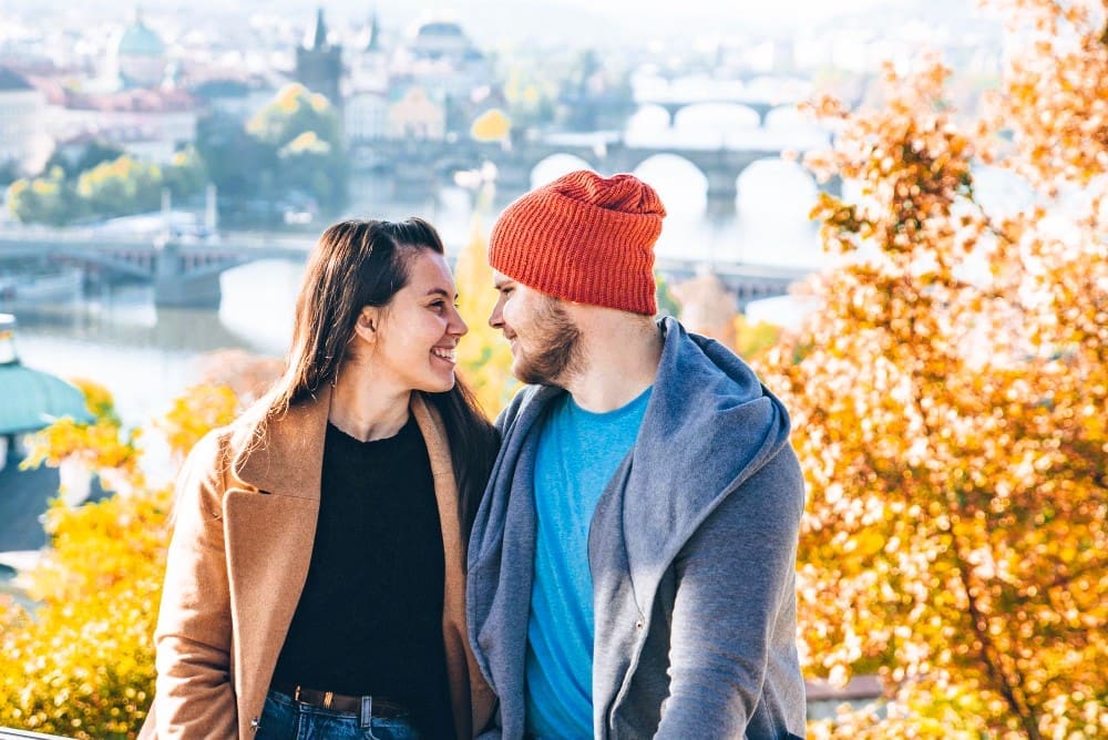 A happy couple smiling at each other, with the city of Prague and its bridges in the blurred background during autumn.