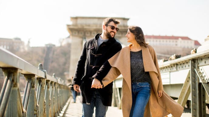 A happy couple holding hands and smiling at each other while walking on the Széchenyi Chain Bridge in Budapest.