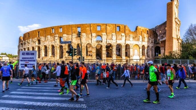 A large crowd of runners and spectators walk past the iconic Roman Colosseum on a sunny day, with a sign indicating "42.195 KM" and "RUNNER ENTRANCE MARATHON START."