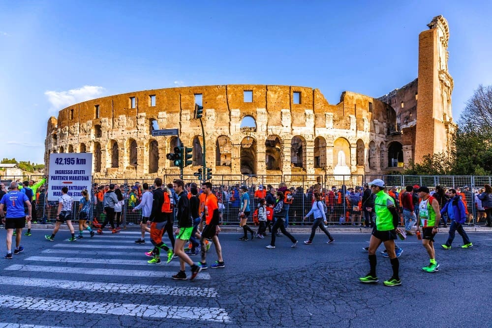 A large crowd of runners and spectators walk past the iconic Roman Colosseum on a sunny day, with a sign indicating "42.195 KM" and "RUNNER ENTRANCE MARATHON START."