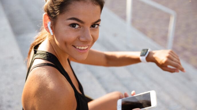 A smiling female runner with wireless earbuds and a smartwatch, holding a smartphone, looking at the camera.