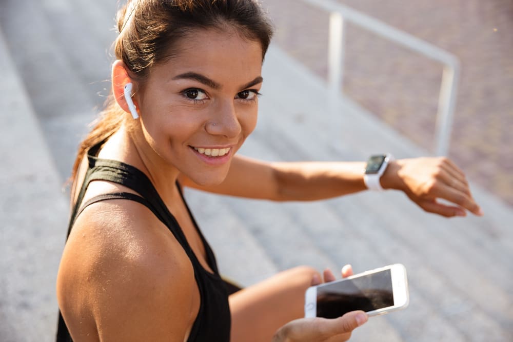 A smiling female runner with wireless earbuds and a smartwatch, holding a smartphone, looking at the camera.