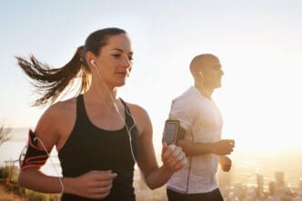 A woman in a black tank top and a man in a white t-shirt are running outdoors, backlit by bright sunlight. Both wear earphones and smartphone armbands, appearing focused on their workout.
