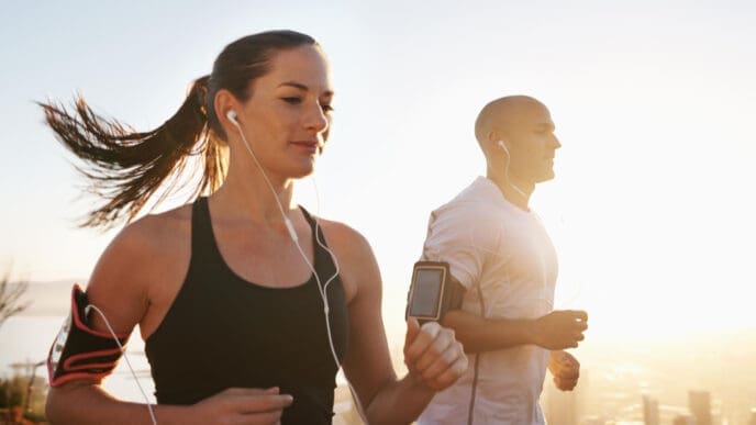 A woman in a black tank top and a man in a white t-shirt are running outdoors, backlit by bright sunlight. Both wear earphones and smartphone armbands, appearing focused on their workout.