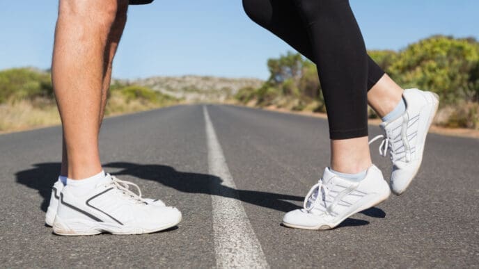 Close-up of two pairs of feet in running shoes, standing on a road with a white line, facing each other.