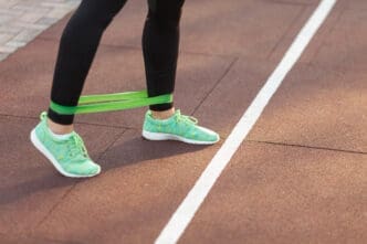A close-up of a person's legs and feet wearing black leggings and green athletic shoes, with a green resistance band looped around their ankles, performing an exercise on a running track.