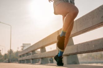 Close-up of a runner's legs and feet in black running shoes and colorful socks, in mid-stride on a paved surface with a concrete railing, under bright sunlight.