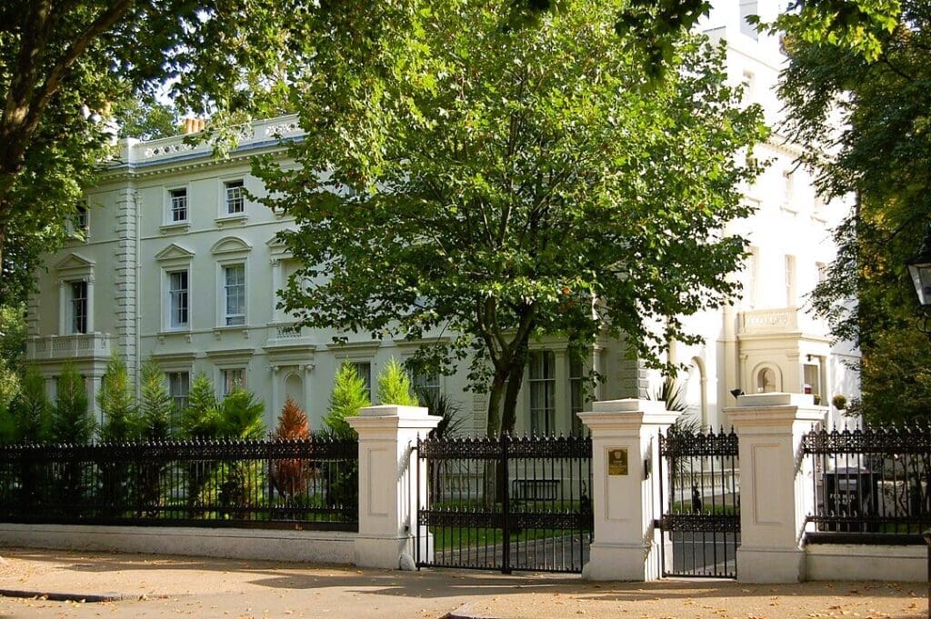 The grand, white facade of 24 Kensington Palace Gardens (Russian Embassy) in London, behind black gates and mature green trees.