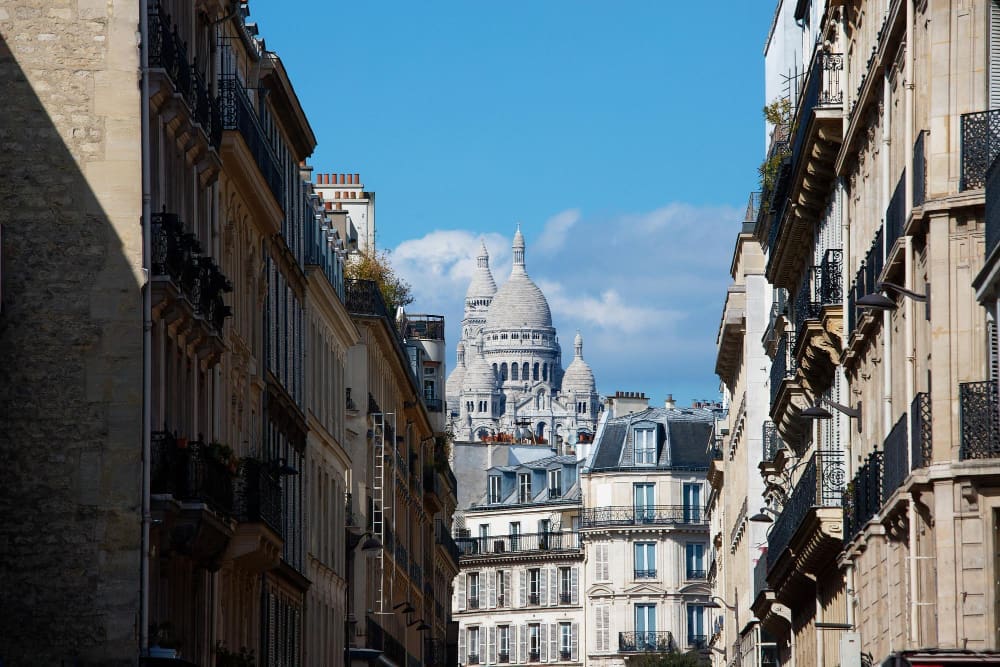 A narrow Parisian street lined with classic Haussmann-style buildings leads towards the iconic white domes of the Sacré-Cœur Basilica in Montmartre, visible in the distance under a blue sky.
