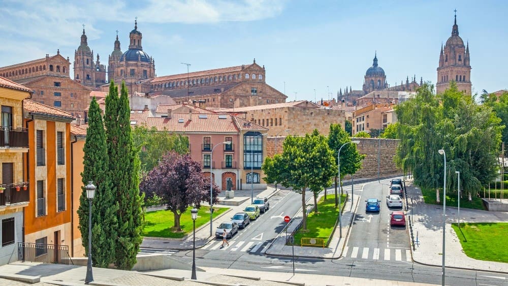 A panoramic view of Salamanca, Spain, showing a city street with cars and trees in the foreground, leading towards historical stone buildings with red roofs, and the prominent spires and domes of its famous cathedrals under a clear blue sky.