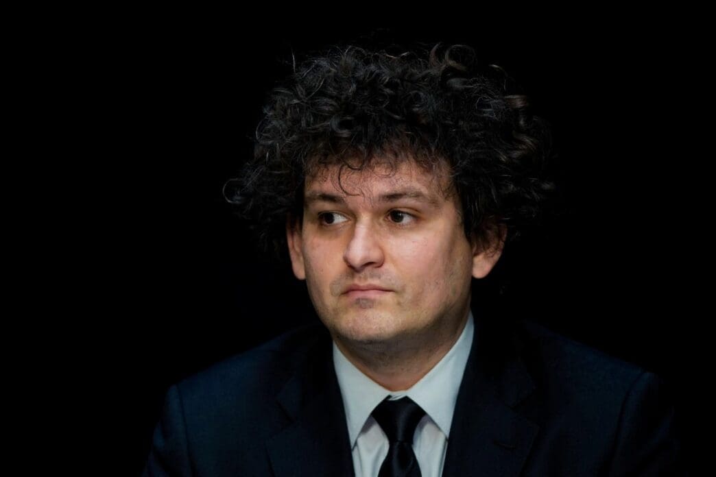 A close-up portrait of Sam Bankman-Fried, with curly dark hair, wearing a dark suit and tie, looking off to his left with a neutral expression against a dark background.
