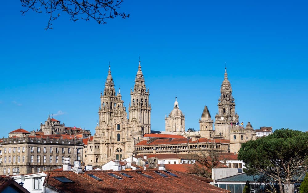A panoramic view of the Santiago de Compostela Cathedral, with its ornate Baroque facades and multiple spires, rising above the red-tiled roofs of the city, under a clear blue sky.