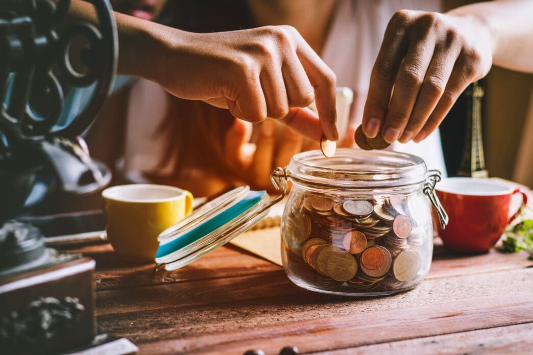 A close-up of a person's hands dropping coins into a glass jar filled with various currencies, symbolizing saving money, on a wooden table with other household items.