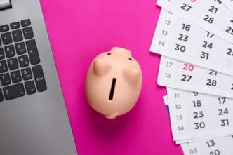 A small piggy bank next to a laptop keyboard and scattered calendar pages on a bright pink background.