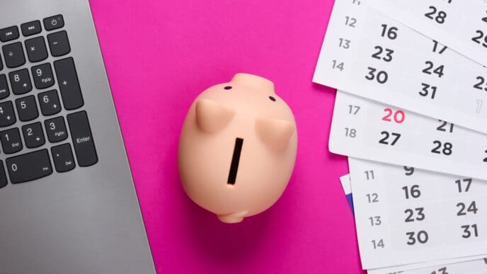 A small piggy bank next to a laptop keyboard and scattered calendar pages on a bright pink background.