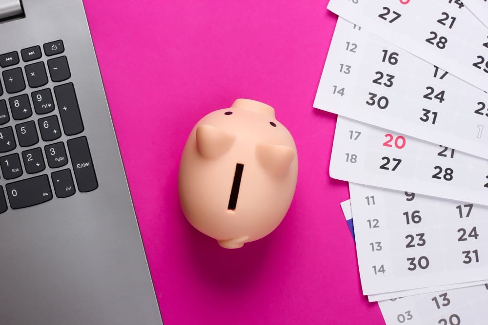 A small piggy bank next to a laptop keyboard and scattered calendar pages on a bright pink background.