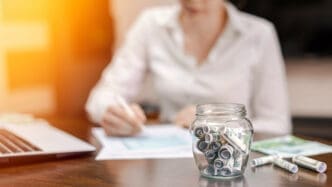 A glass jar filled with rolled-up US dollar bills sits on a wooden desk, with a person writing in the blurred background, next to a laptop and papers, symbolizing savings and financial planning.