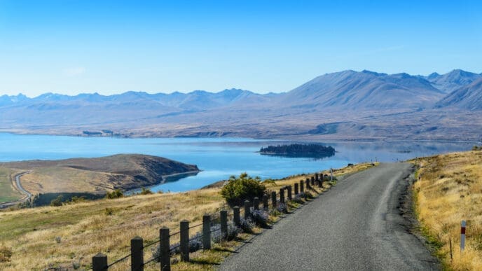 A scenic landscape view of a winding road leading towards a large blue lake with a small, forested island, surrounded by dry golden hills and distant mountains under a clear blue sky, in New Zealand.