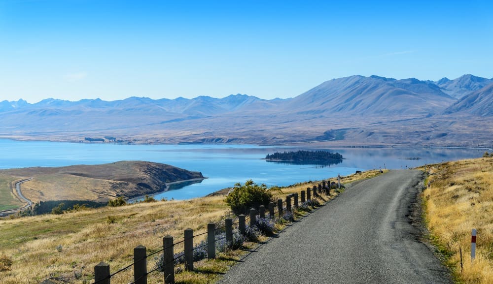 A scenic landscape view of a winding road leading towards a large blue lake with a small, forested island, surrounded by dry golden hills and distant mountains under a clear blue sky, in New Zealand.
