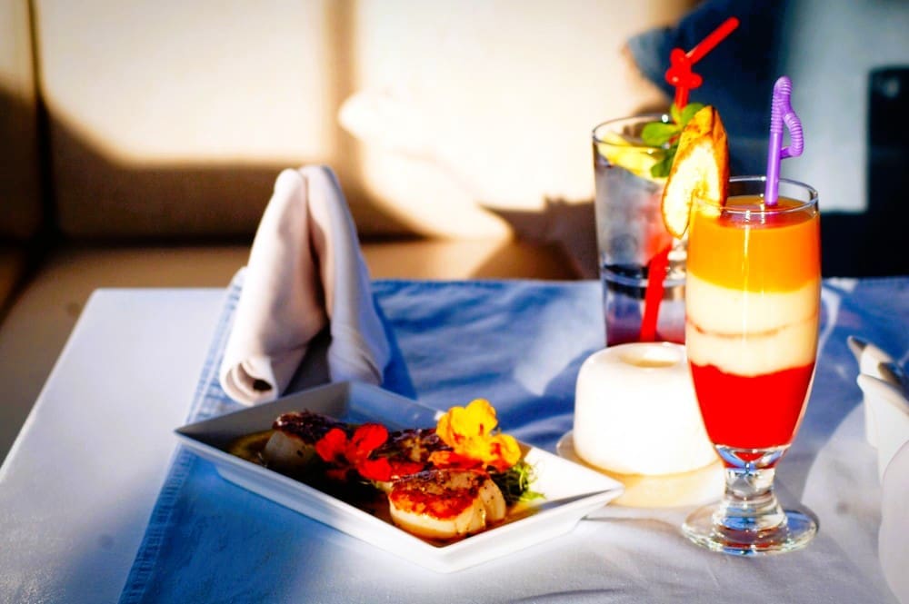 A beautifully presented meal with two drinks on a table, including a layered red, white, and orange cocktail and a plate with seared scallops garnished with edible flowers.