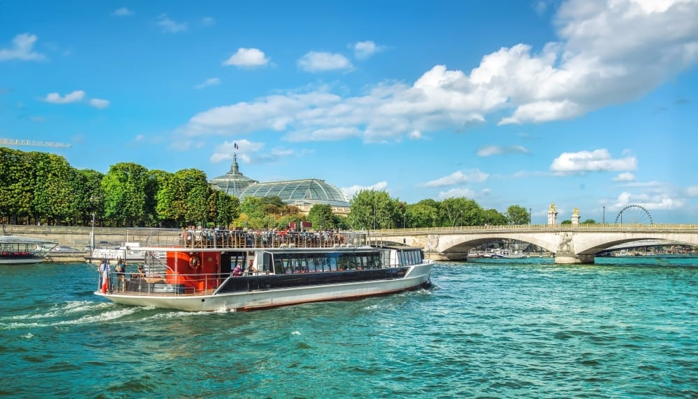 A large tourist boat filled with passengers cruises on the wide Seine River in Paris, with trees, a bridge, and a glass-domed building visible in the background under a partly cloudy sky.