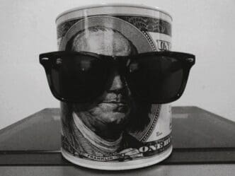 A black and white image of a cylindrical coin bank with a depiction of Benjamin Franklin from a hundred-dollar bill, wearing dark sunglasses, standing on a dark surface.