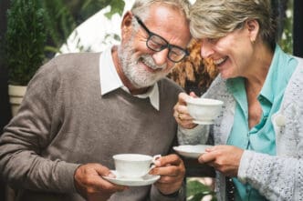 A happy senior couple with white hair and glasses share a joyful moment over coffee or tea, smiling and looking down, suggesting a pleasant conversation in a cozy setting.