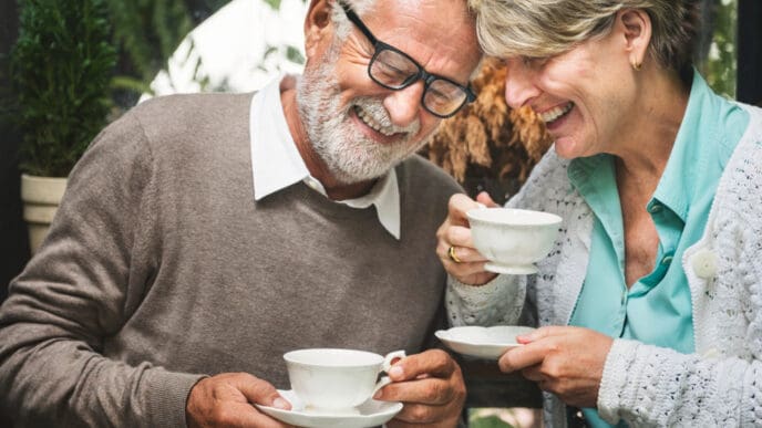 A happy senior couple with white hair and glasses share a joyful moment over coffee or tea, smiling and looking down, suggesting a pleasant conversation in a cozy setting.