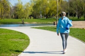 Rear view of a woman in activewear walking on a curved path in a park, carrying a yoga mat.