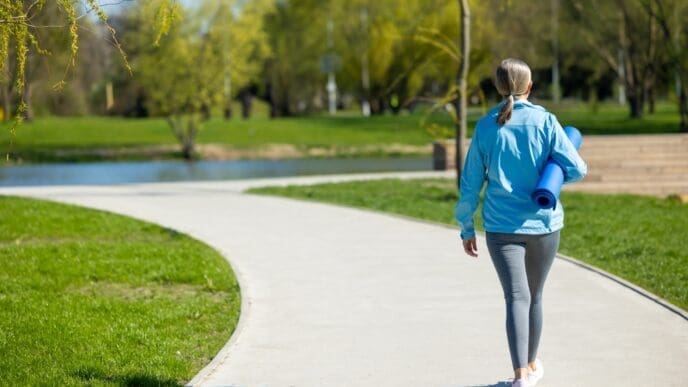 Rear view of a woman in activewear walking on a curved path in a park, carrying a yoga mat.