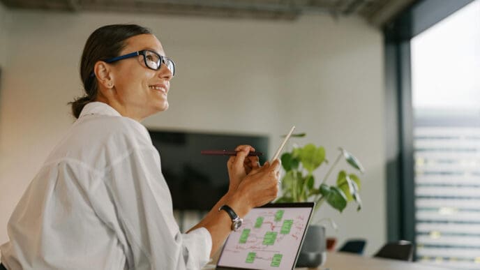 A smiling businesswoman in glasses looking up while working on a laptop and holding a pen in a modern office.