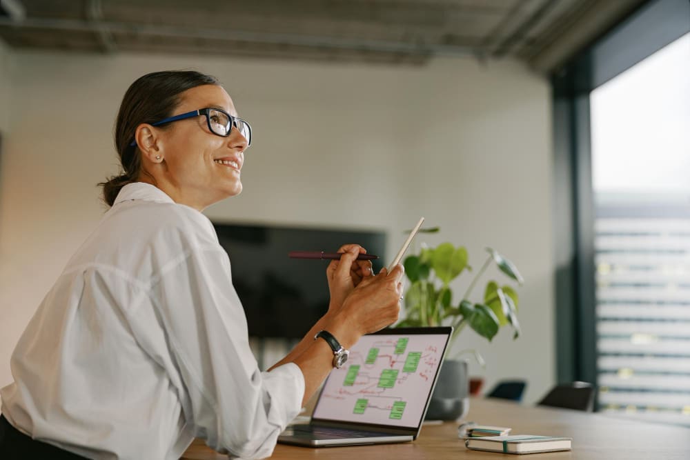 A smiling businesswoman in glasses looking up while working on a laptop and holding a pen in a modern office.