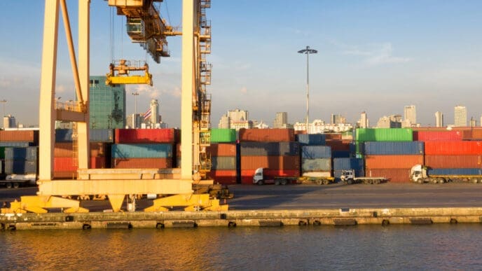 A large yellow gantry crane and stacks of colorful shipping containers line a port, with several trucks and a distant city skyline in the background, under a blue sky.
