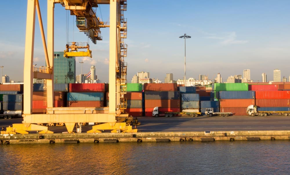 A large yellow gantry crane and stacks of colorful shipping containers line a port, with several trucks and a distant city skyline in the background, under a blue sky.