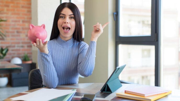 An excited young woman holding a piggy bank and pointing upwards, with a tablet and papers on a desk in front of her.
