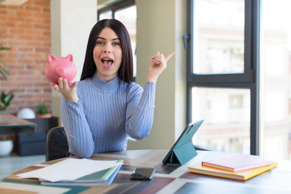 An excited young woman holding a piggy bank and pointing upwards, with a tablet and papers on a desk in front of her.
