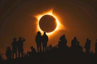 Silhouettes of a group of people, with two figures standing prominently in the center, watching a total solar eclipse with a bright, fiery corona around the moon's silhouette against a dark sky.