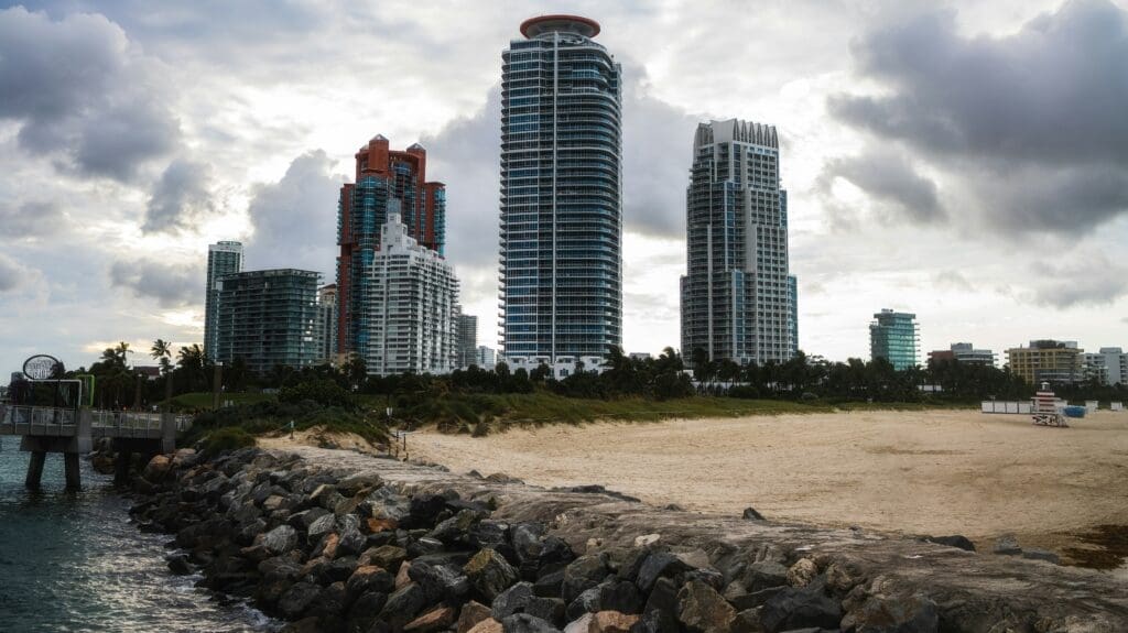 The skyline of South Beach, Miami, with towering high-rises and a sandy beach, seen from the South Pointe Park jetty at sunset under a cloudy sky.