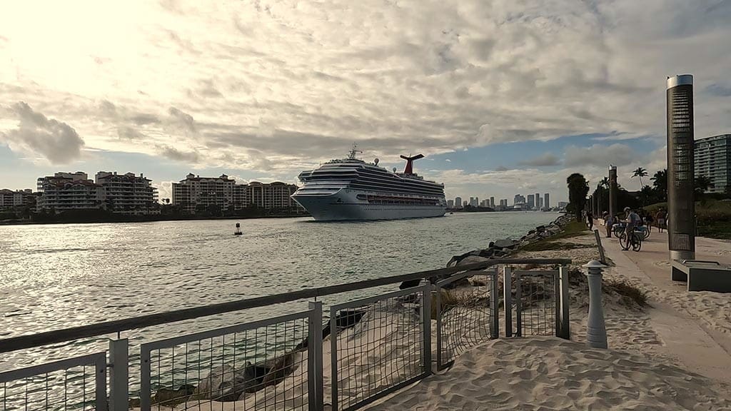 A large Carnival cruise ship sails down a channel towards the ocean, with South Pointe Park's sandy beach, promenade, and residential buildings in the foreground and a distant city skyline under a cloudy sky at sunset.