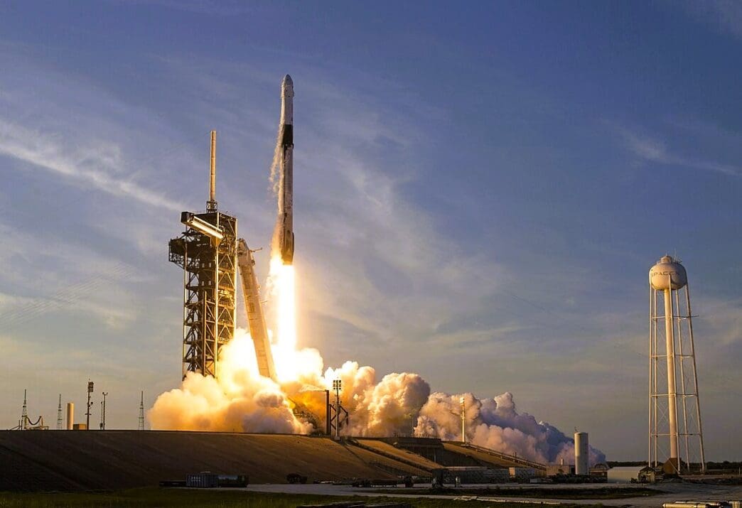 A SpaceX Falcon 9 rocket launching from Kennedy Space Center, with a fiery exhaust plume and thick smoke, against a clear sky.