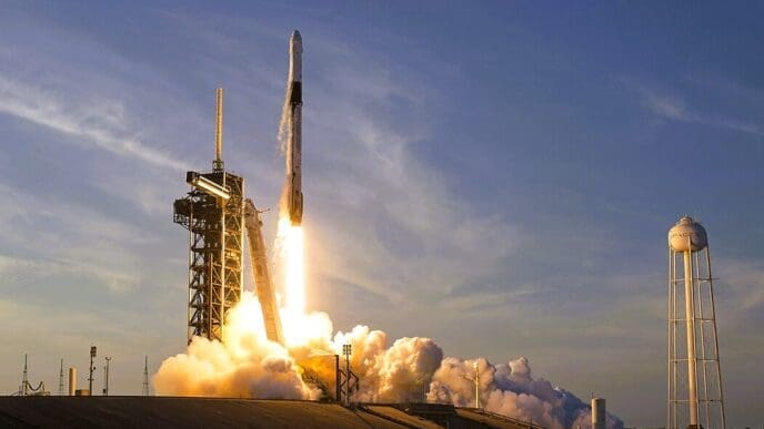 A SpaceX Falcon 9 rocket launching from Kennedy Space Center, with a fiery exhaust plume and thick smoke, against a clear sky.