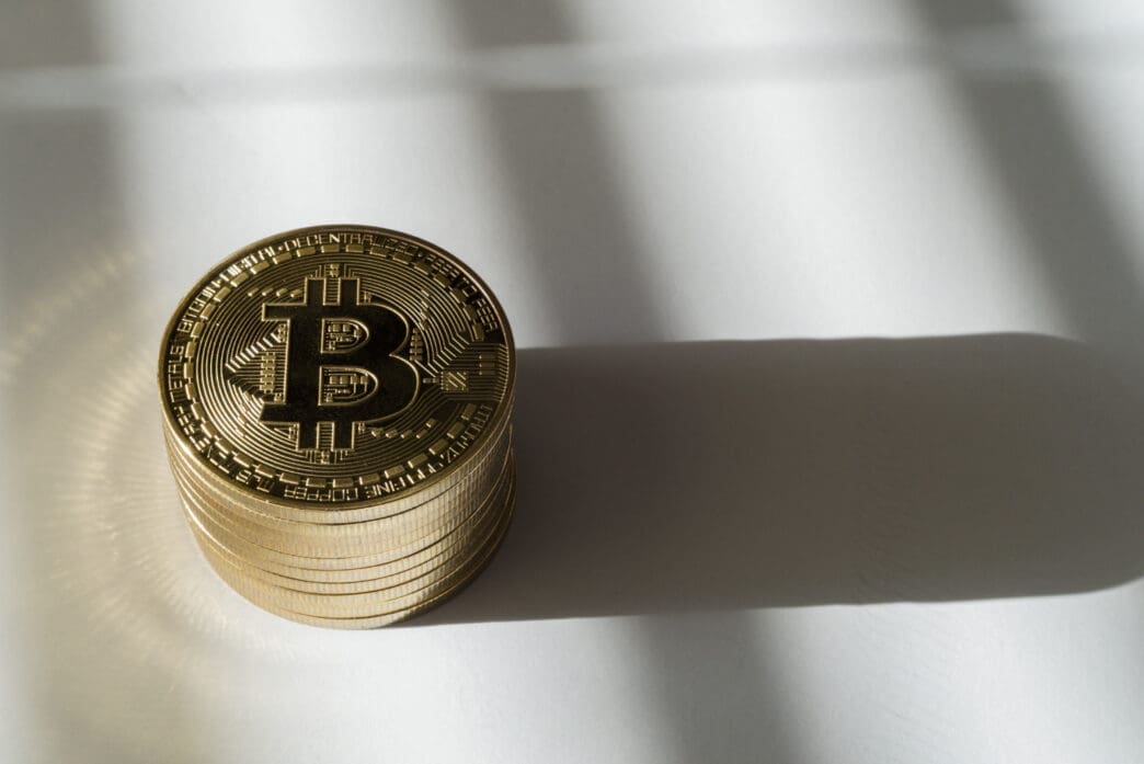 A high-angle view of a neat stack of golden Bitcoin coins on a plain white surface, casting a distinct, elongated shadow.